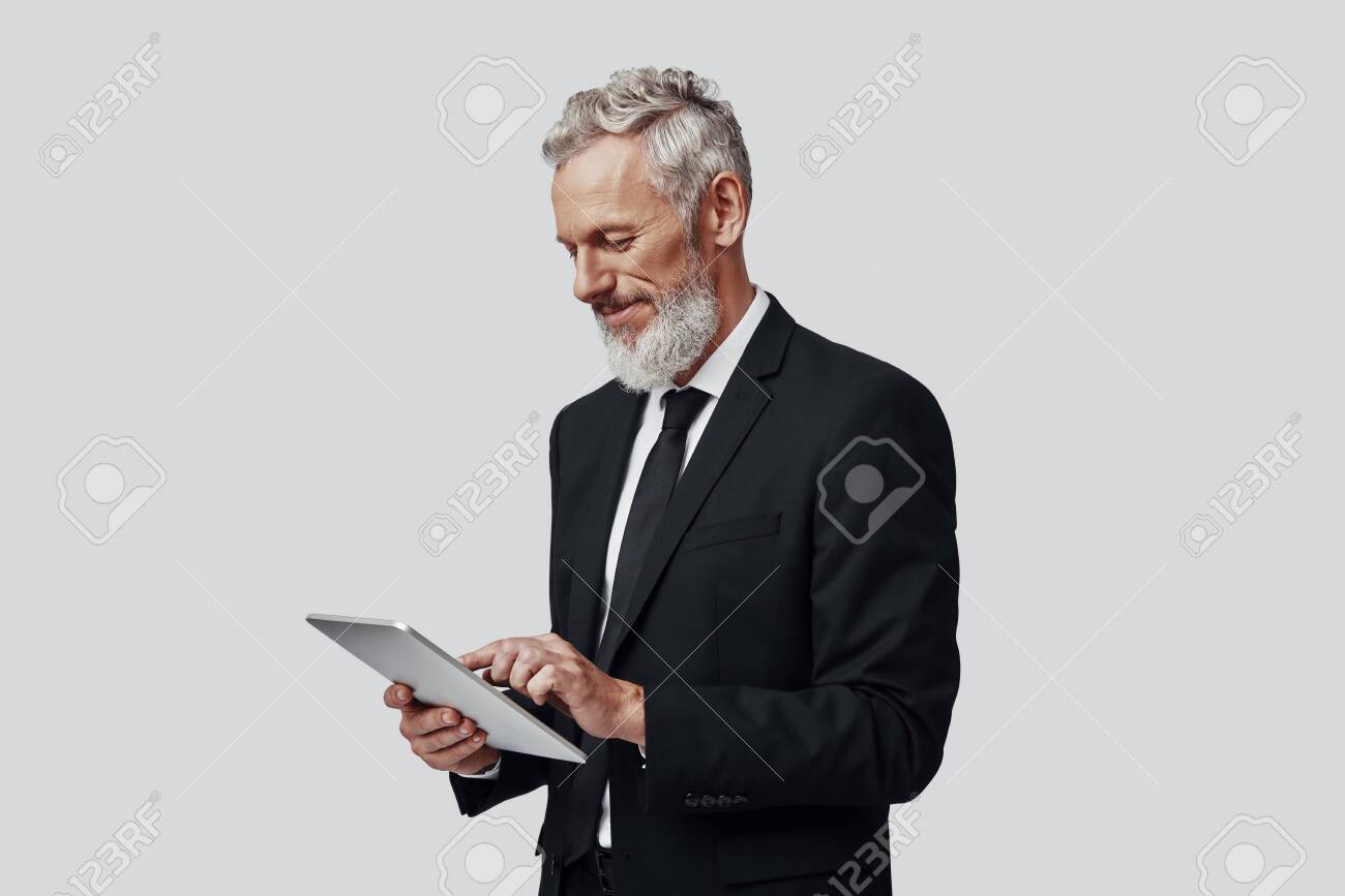 Thoughtful mature man in full suit working using digital tablet and smiling while standing against grey background - 137917279 Thoughtful mature man in full suit working using digital tablet and smiling while standing against grey background - 137917279