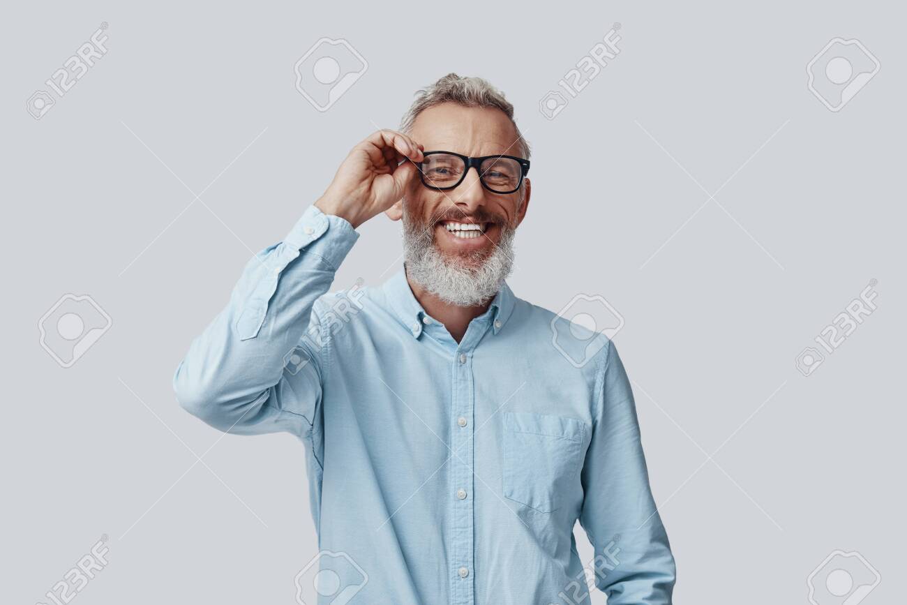 Happy mature man looking at camera and adjusting eyewear while standing against grey background - 138444956 Happy mature man looking at camera and adjusting eyewear while standing against grey background - 138444956