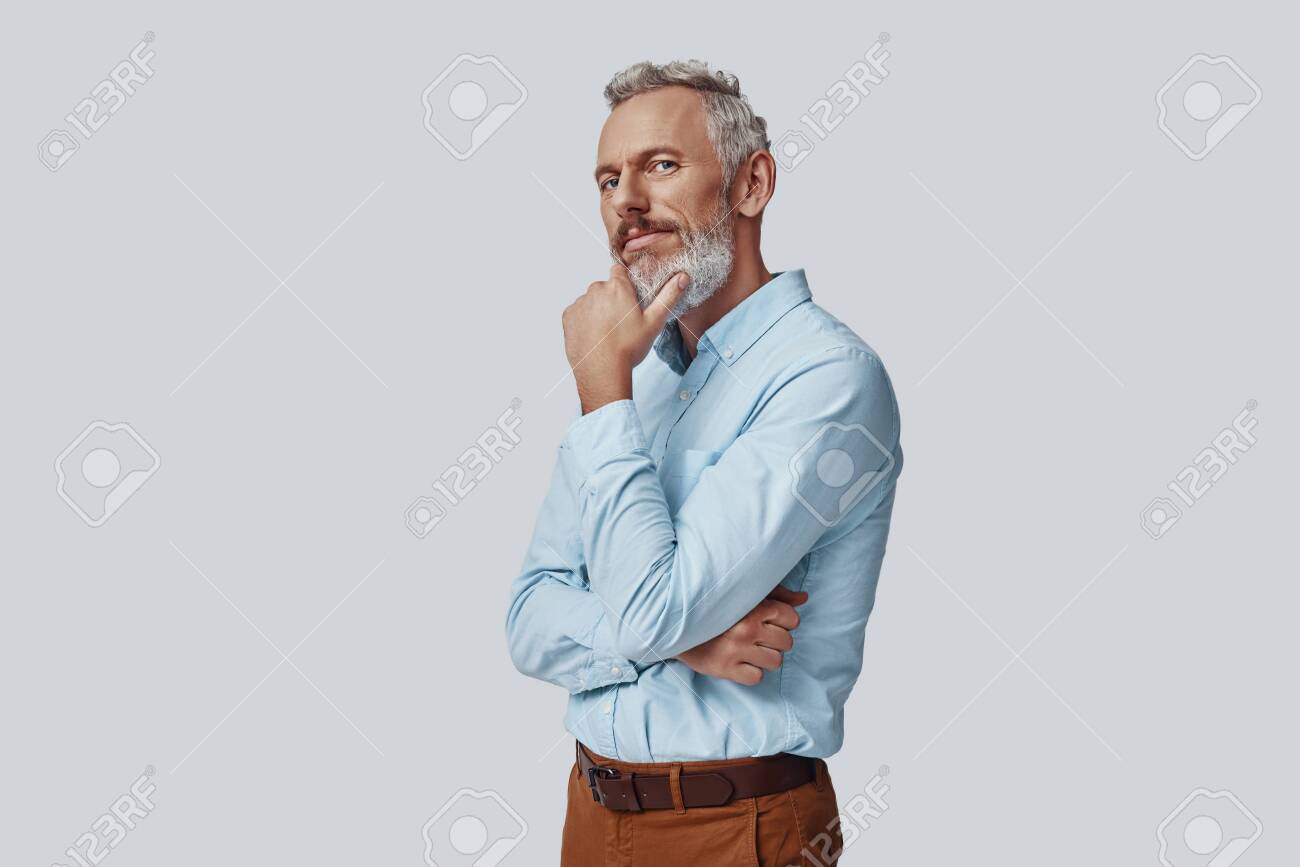 Thoughtful mature man looking at camera and keeping hand on chin while standing against grey background - 138445034 Thoughtful mature man looking at camera and keeping hand on chin while standing against grey background - 138445034
