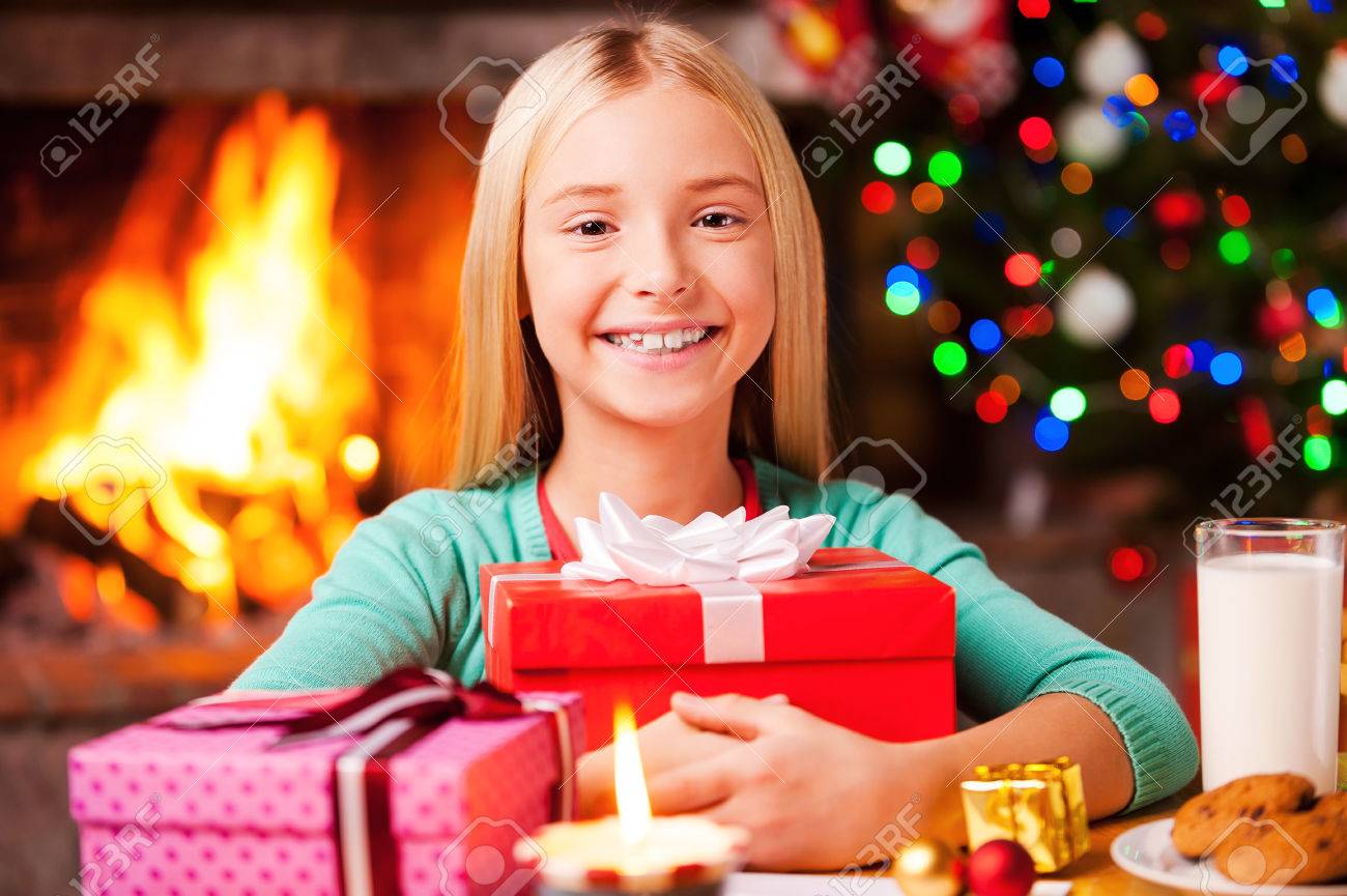 私のクリスマス プレゼント ギフト用の箱を抱きしめるとクリスマス ツリーと背景の暖炉のそば テーブルに座って笑顔かわいい女の子 の写真素材 画像素材 Image