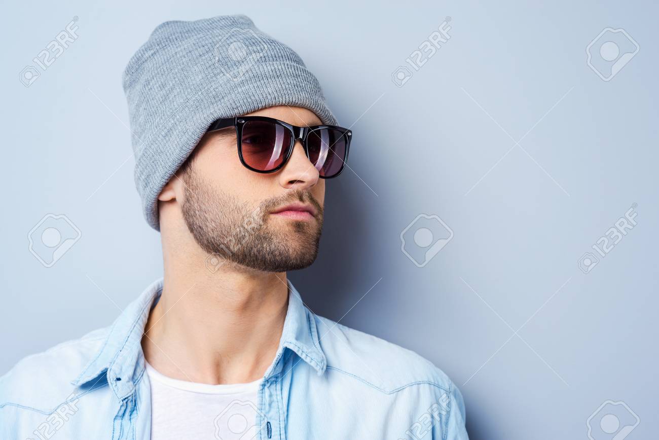 Cool and handsome. Handsome young stylish man in sunglasses and hat looking away while standing against grey background - 37823481 Cool and handsome. Handsome young stylish man in sunglasses and hat looking away while standing against grey background - 37823481
