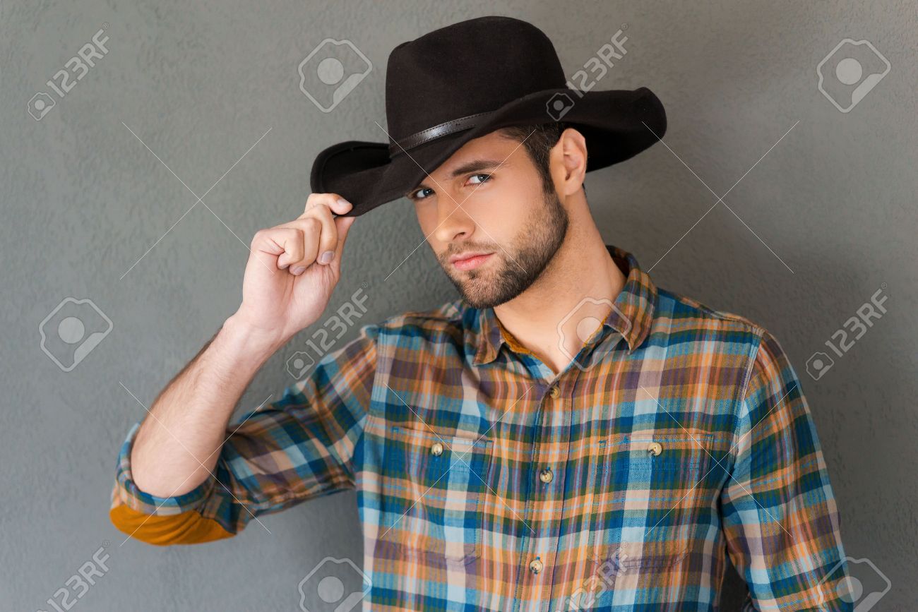 Estilo Vaquero. Hombre Joven Hermoso Que Ajusta Su Sombrero De Vaquero Y Mirando A La Mientras Está Pie El Fondo Gris Fotos, Retratos, Imágenes Y Fotografía De Archivo Libres