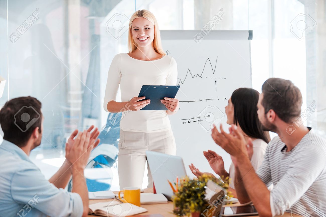 Great presentation! Cheerful young woman standing near whiteboard and smiling while her colleagues sitting at the desk and applauding - 41179606 Great presentation! Cheerful young woman standing near whiteboard and smiling while her colleagues sitting at the desk and applauding - 41179606