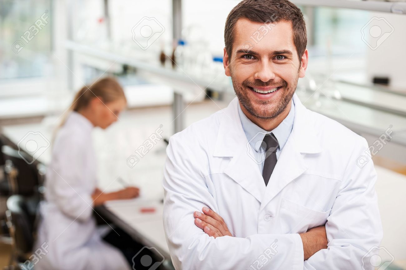 Confident Scientist. Happy Young Male Scientist Keeping Arms Crossed And Looking At Camera While His Female Colleague Working In The Background Stock Photo, Picture And Royalty Free Image. Image 44278176.