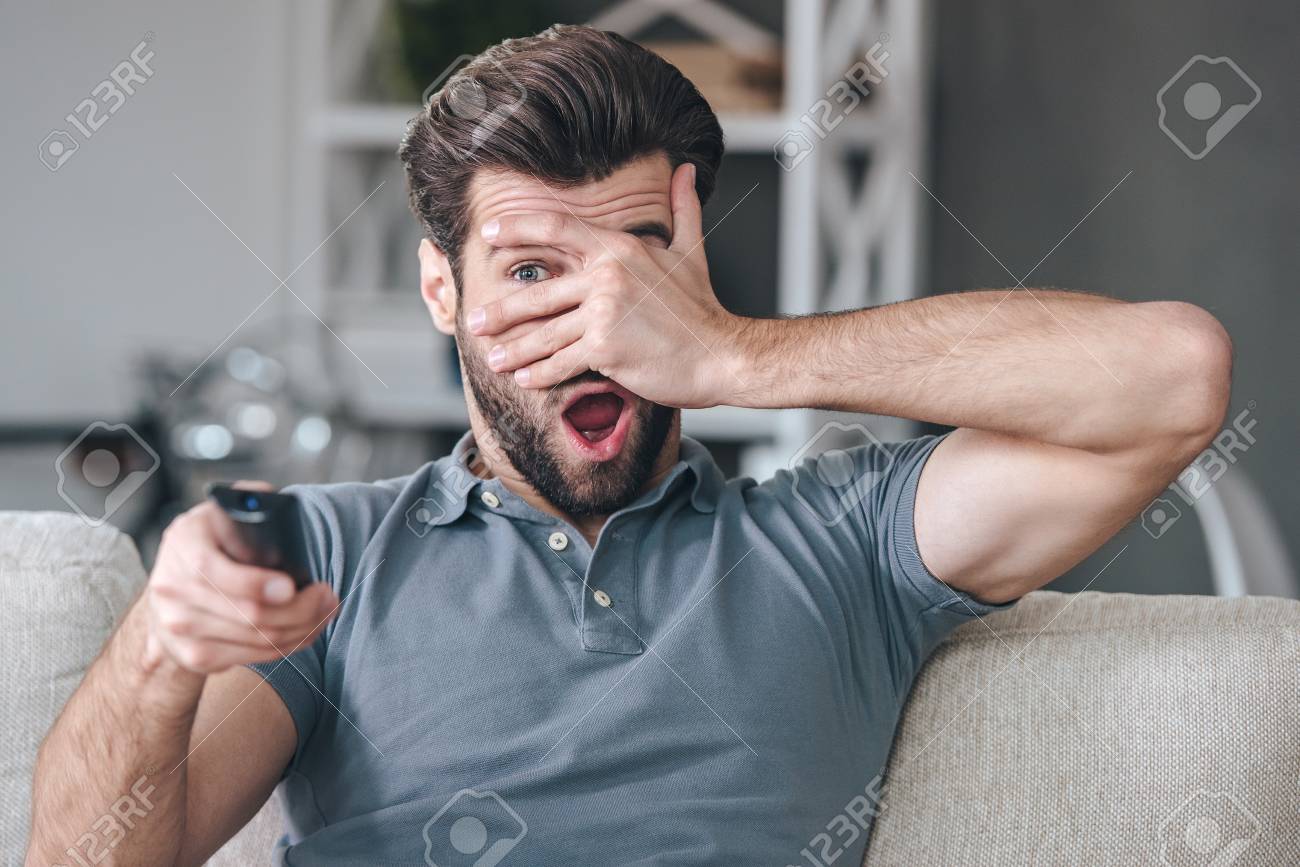 58677531-horror-movie-handsome-young-man-holding-remote-control-and-watching-tv-through-his-fingers-while-sit.jpg