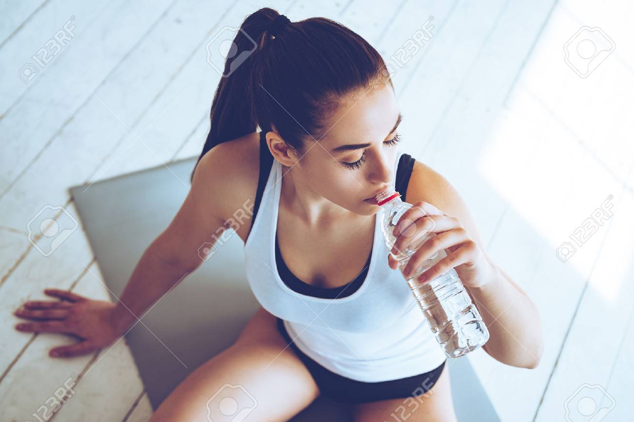 Sip of fresh water after great workout. Top view of beautiful young woman drinking water while sitting on exercise mat at gym - 55095848 Sip of fresh water after great workout. Top view of beautiful young woman drinking water while sitting on exercise mat at gym - 55095848