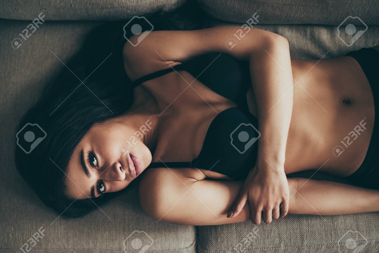 Belleza En Lencería. Vista Superior De La Joven Mujer De Raza Mixta En Ropa Interior Negro Mirando A Cámara Mientras Se Está Acostado En El Sofá Fotos, Retratos, Imágenes Y Fotografía