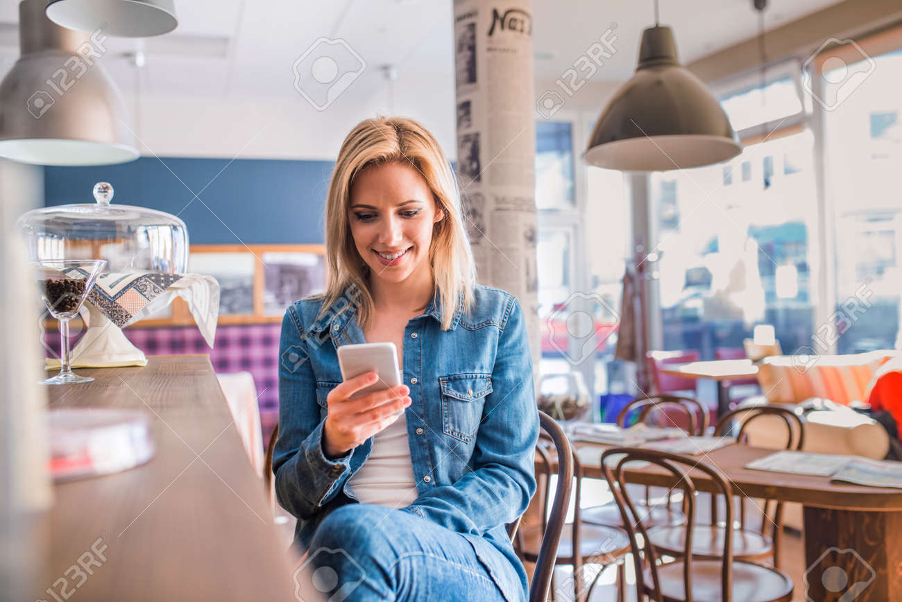 Blond woman in denim shirt sitting at the bar in modern city cafe, holding a smart phone, texting - 56194129 Blond woman in denim shirt sitting at the bar in modern city cafe, holding a smart phone, texting - 56194129