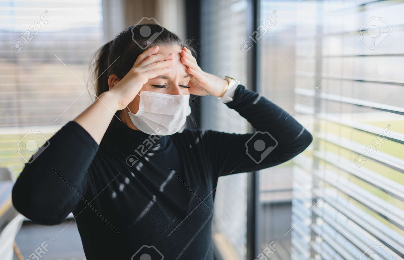 Woman with headache and face masks indoors at home, Corona virus concept. - 144720744 Woman with headache and face masks indoors at home, Corona virus concept. - 144720744