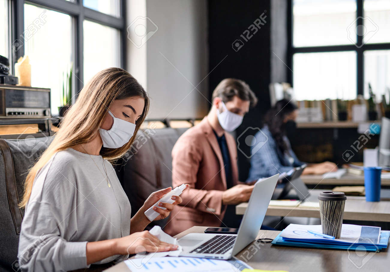 Portrait of young businesspeople with face masks working indoors in office, disinfecting laptop. - 155417596 Portrait of young businesspeople with face masks working indoors in office, disinfecting laptop. - 155417596