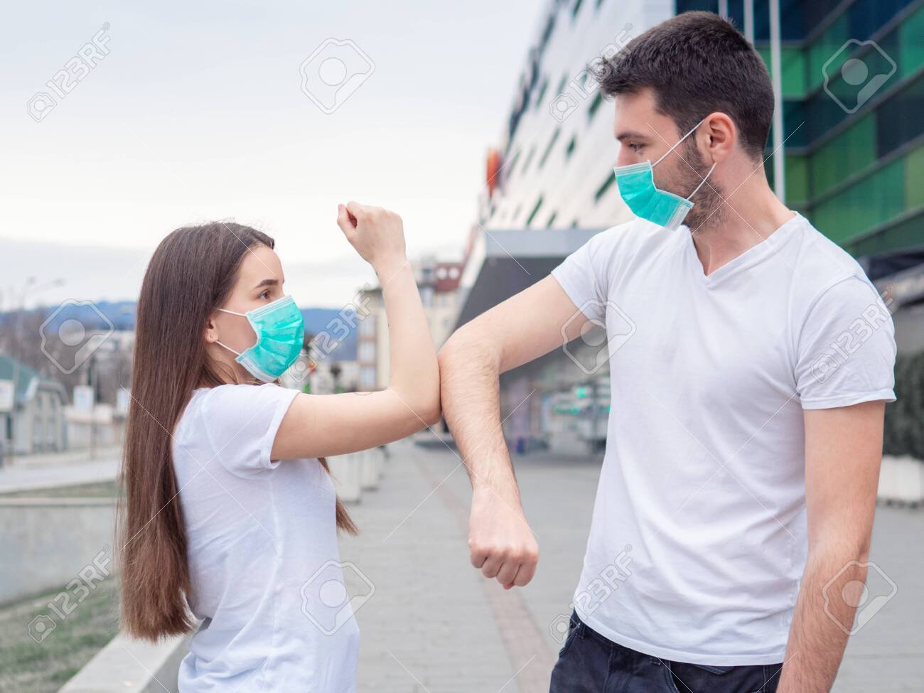 Two people woman and man wearing medical mask, friends greet each other with their elbows, instead of shaking hands. Elbow bump. A new greeting way to avoid the spread of coronavirus. - 143409686 Two people woman and man wearing medical mask, friends greet each other with their elbows, instead of shaking hands. Elbow bump. A new greeting way to avoid the spread of coronavirus. - 143409686