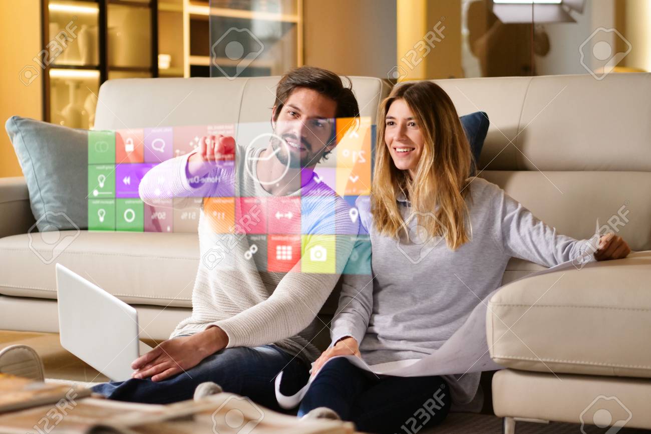 A couple sitting on the sofa controls all the functions of the house such as wi-fi, heating, lighting, and television through holography. Concept of, home automation, automations, future, technology. - 114885931 A couple sitting on the sofa controls all the functions of the house such as wi-fi, heating, lighting, and television through holography. Concept of, home automation, automations, future, technology. - 114885931