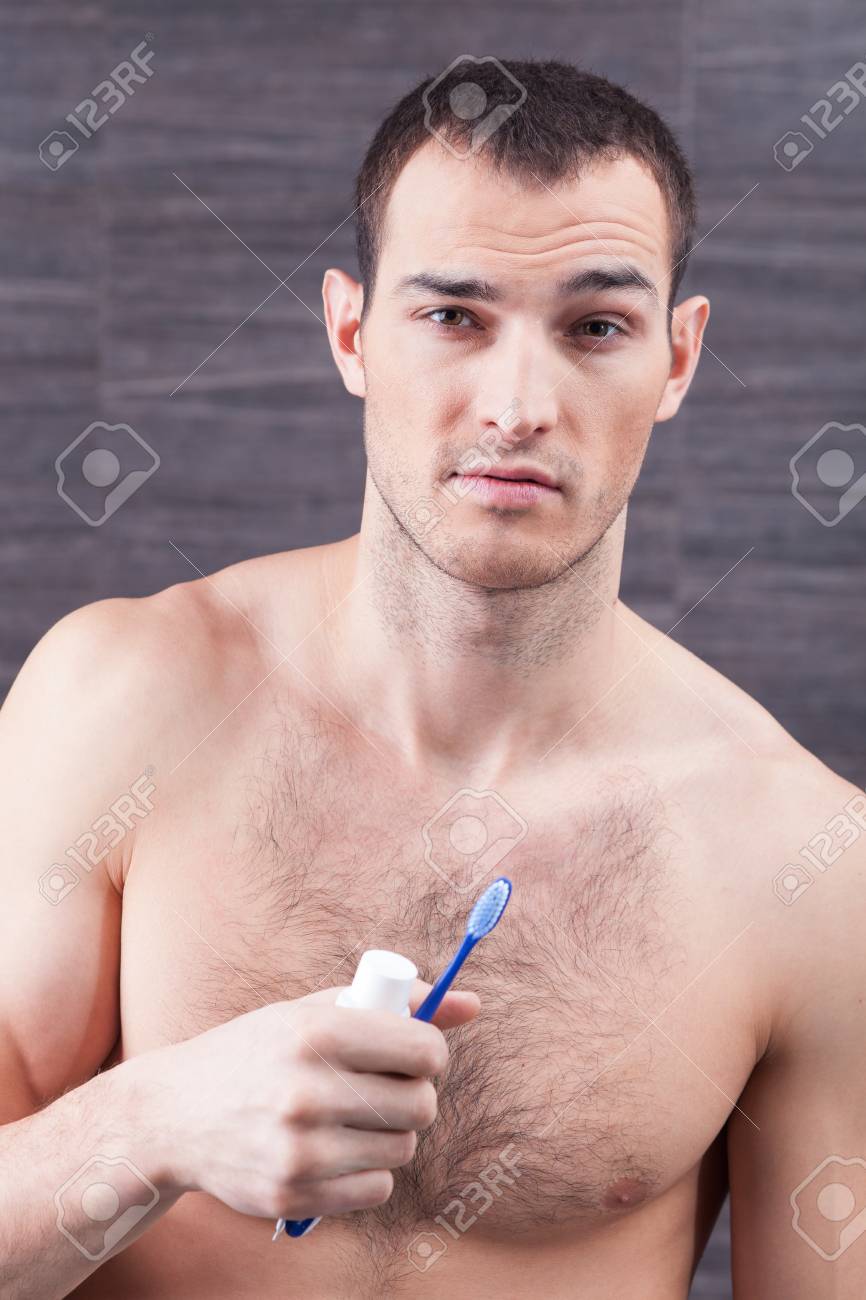 Waist Up Portrait Of Handsome Young Man Preparing For Brushing His Teeth.  He Is Standing In The Bathroom With Naked Torso And Looking Forward  Confidently. The Man Is Holding Brush And Paste