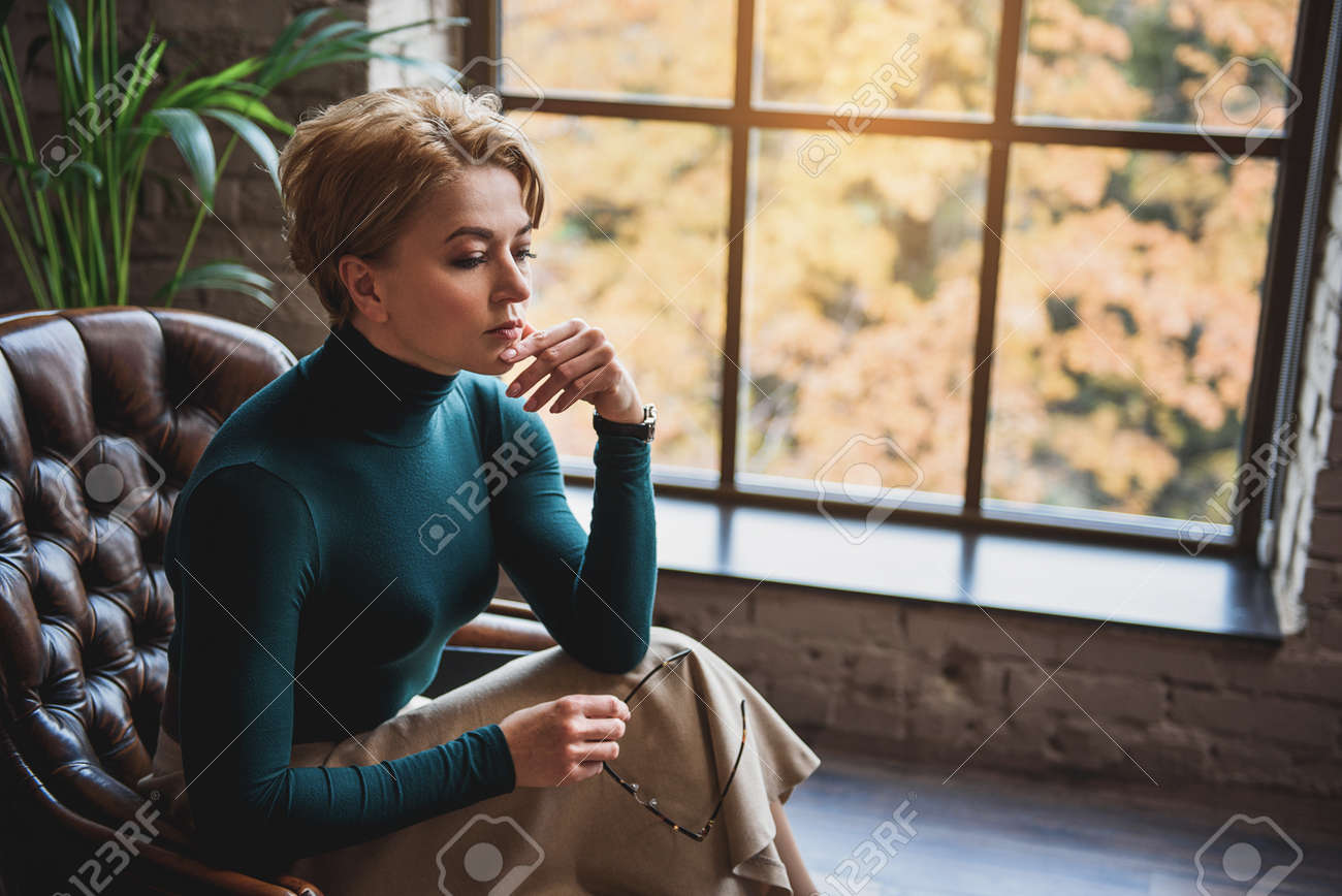 思いやりのある女性は 窓際の椅子に座っています 彼女は床を見て ガラスを保持しています 指で彼女のあごに触れる の写真素材 画像素材 Image