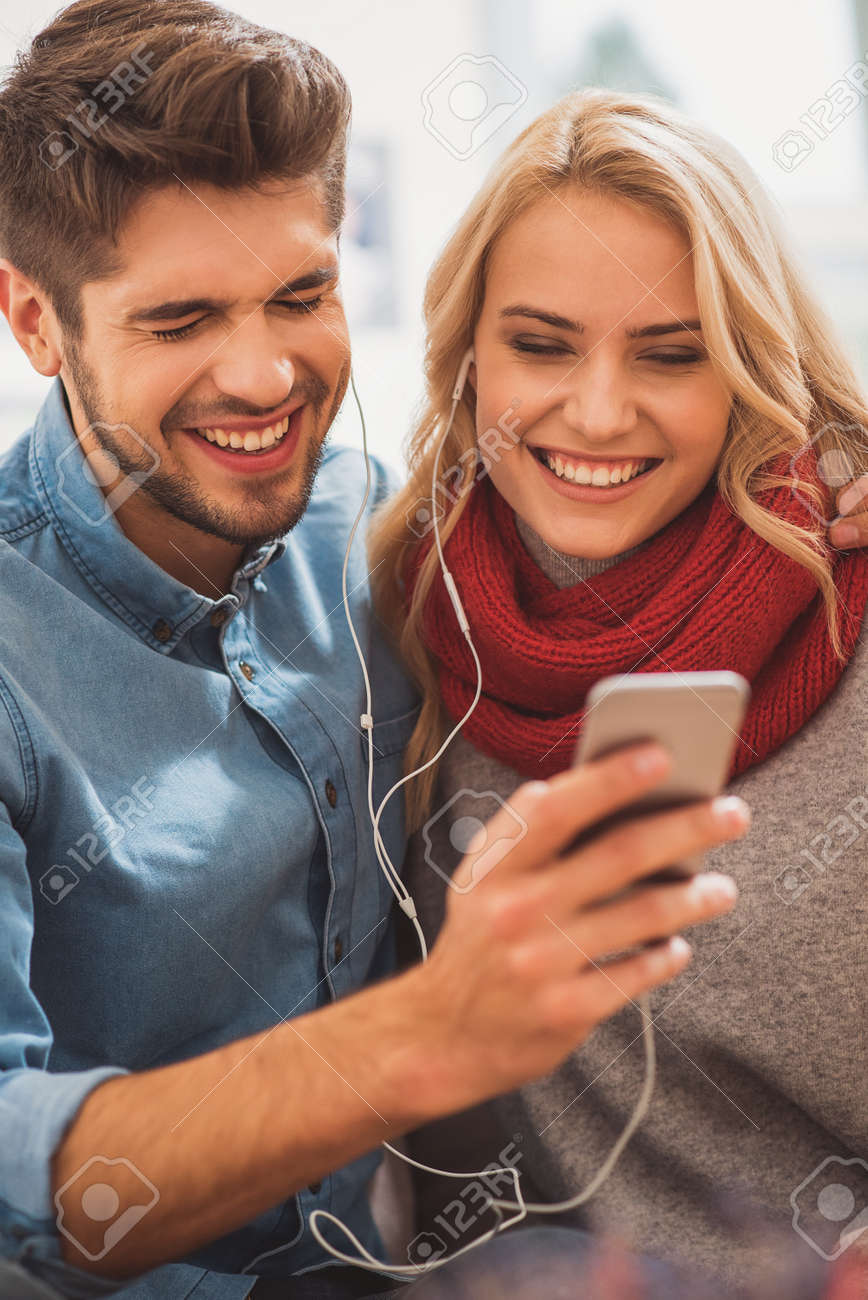 屈託のない夫婦はスマート フォンからおかしい何かを聞いています イヤホンを着て 笑っています 恋人が座っているとハグ の写真素材 画像素材 Image