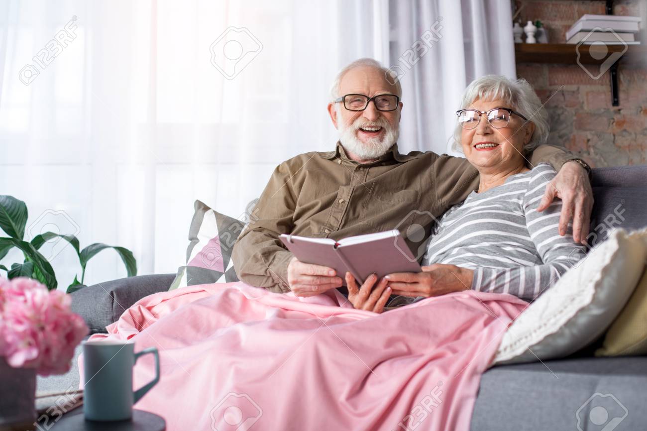 Portrait Of Lovely Married Old Couple Relaxing On Couch With Pillows At  Home. Man Is Hugging Woman While They Are Looking At Camera And Laughing  Stock Photo, Picture and Royalty Free Image.