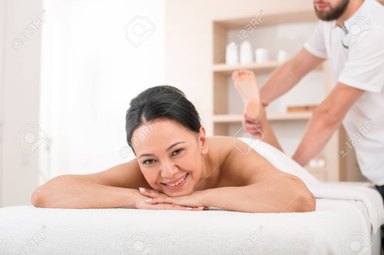 マッサージテーブルの上に横たわり カメラを見ながら笑顔を浮かべた陽気な先輩女性の肖像画 美容院で彼女の足をマッサージセラピスト の写真素材 画像素材 Image