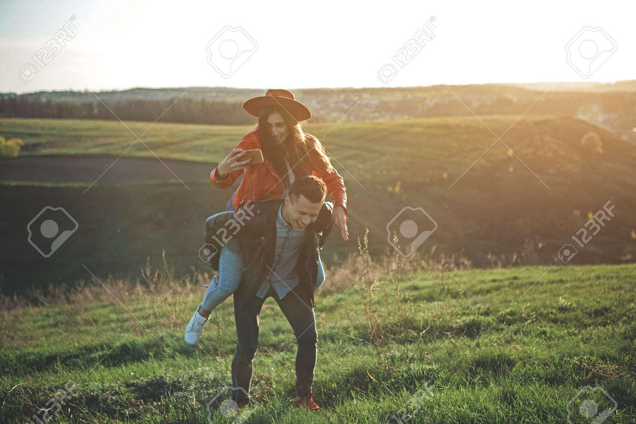 Happy In Love. Full Length Of Full Of Happiness And Joy Couple In Summer  Evening Nature. Boy Is Carrying Girl With Cellphone In Hand. They Are  Smiling And Having Fun Together Stock