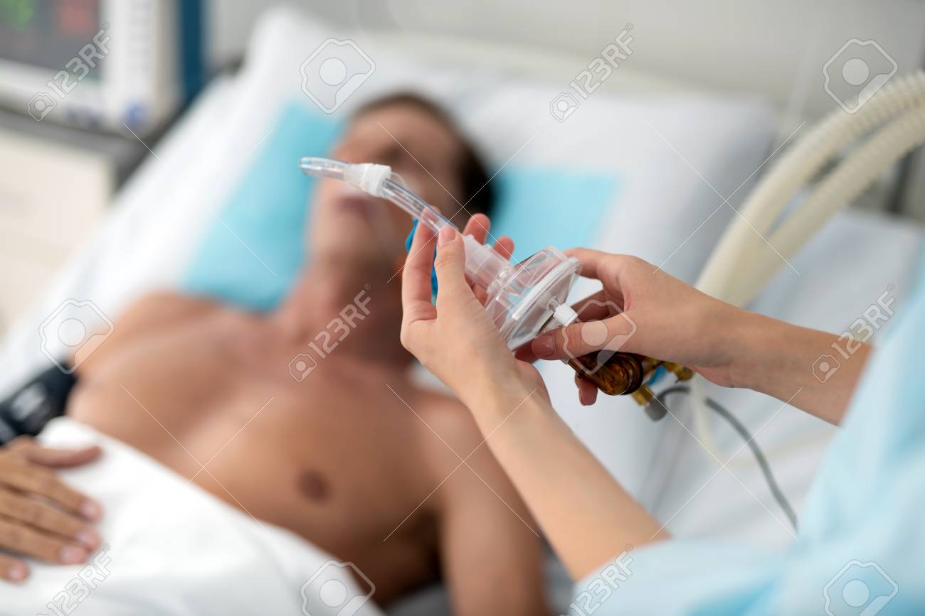 Mechanical ventilation. Close up of female hands preparing tube of breathing machine while middle aged man lying in hospital bed - 109482709 Mechanical ventilation. Close up of female hands preparing tube of breathing machine while middle aged man lying in hospital bed - 109482709