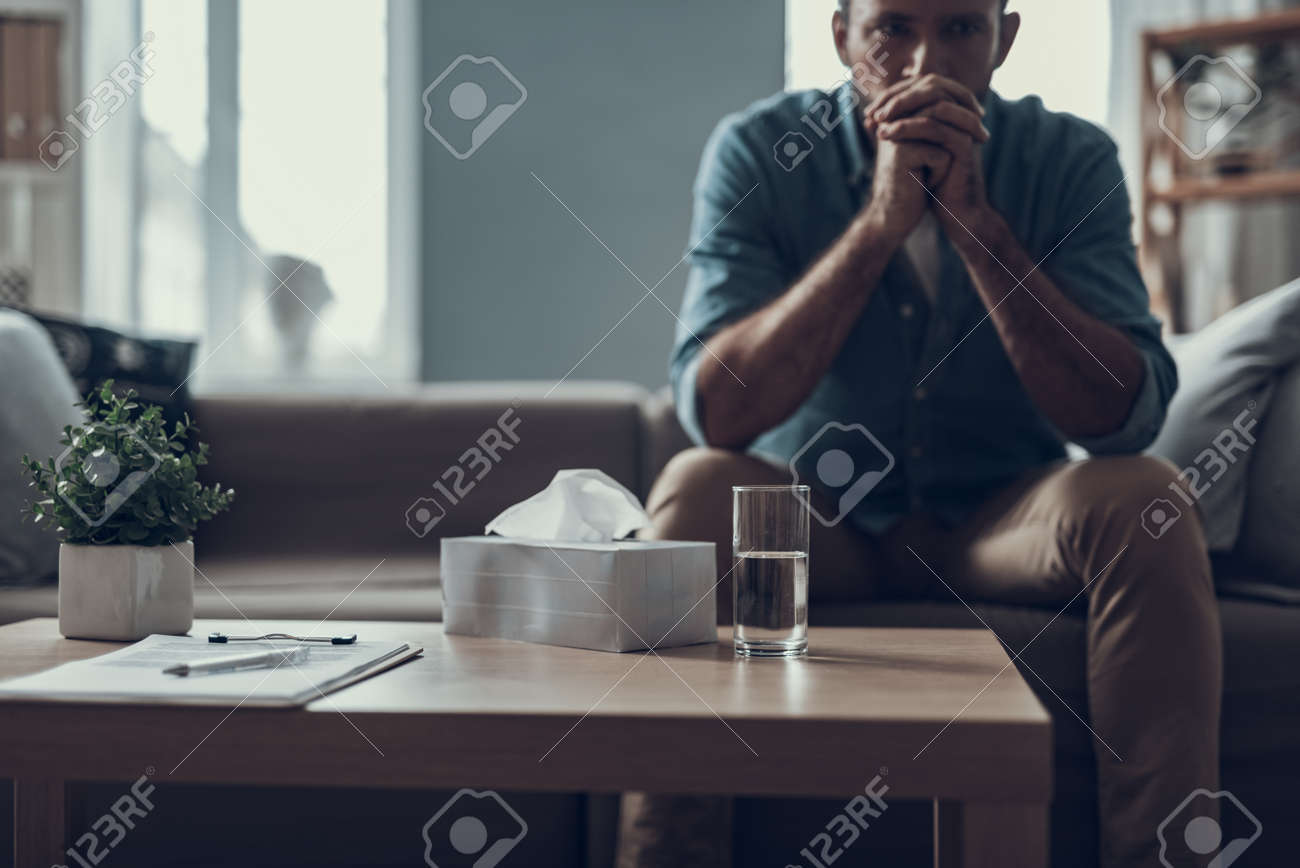 Man sitting on the sofa with his fingers crossed in front of his face and having a glass of water - 114993879 Man sitting on the sofa with his fingers crossed in front of his face and having a glass of water - 114993879
