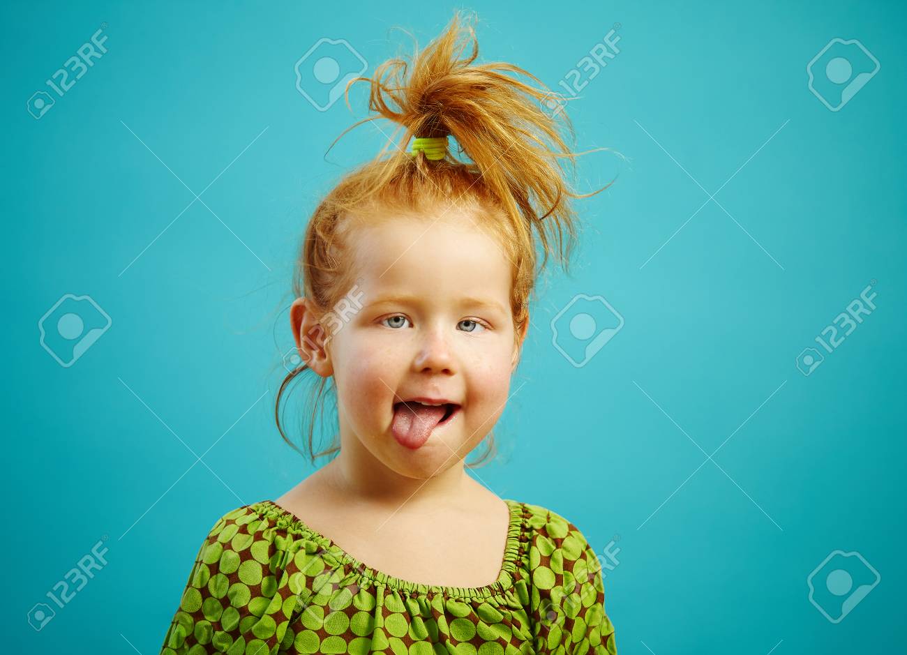 Close-up Shot Of Beautiful Squint Girl Three Years Old With Tongue Out, Has  Red Hair, Is Dressed In Green Dress, Expresses A Sweet Sincerity, Over Blue  Isolated. Funny Baby Licking Her Lips., image size:1300x939