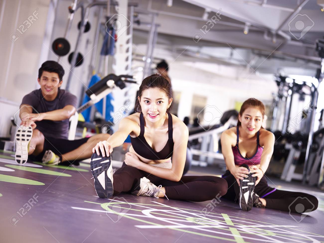 three young asian adult people exercising in gym stretching legs. - 87617765 three young asian adult people exercising in gym stretching legs. - 87617765