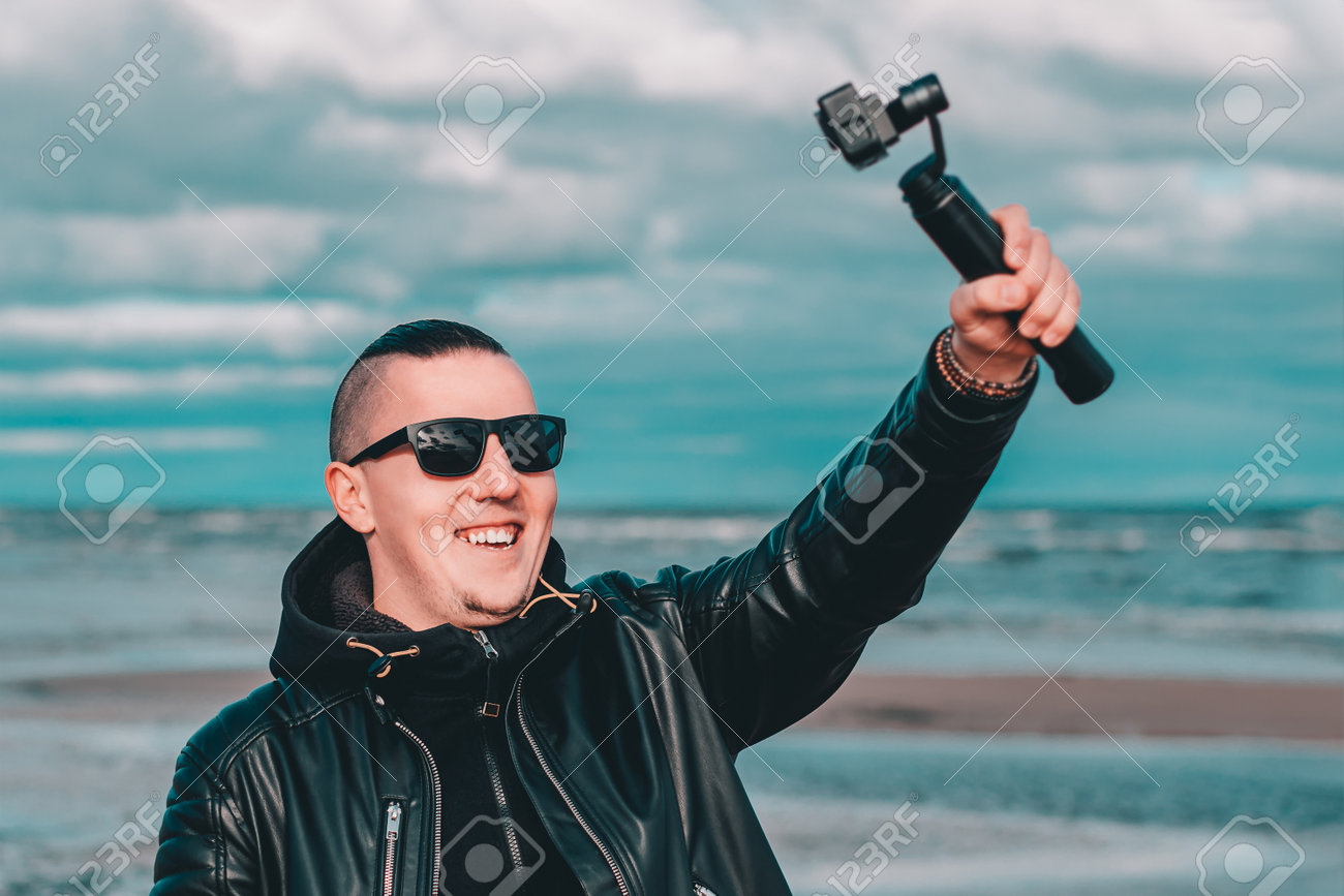 Smiling Young Male Blogger In Sunglasses Making Selfie Or Streaming Video  At The Beach Using Action Camera With Gimbal Camera Stabilizer. Hipster In  Black Clothes Making Photo Against The Sea Фотография, картинки,