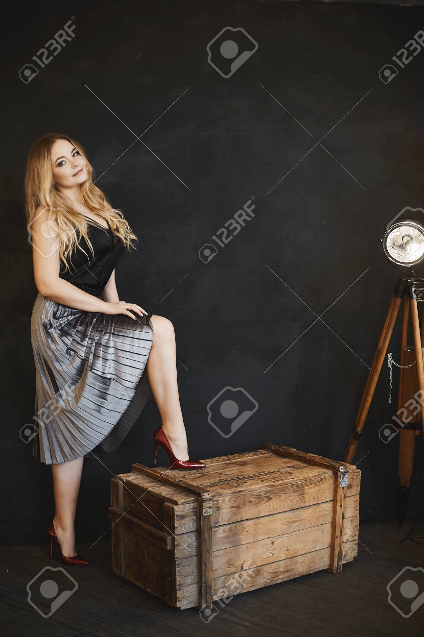 Beautiful and fashionable blonde model girl with big in silver skirt, in black satin blouse and dark-red shoes, stands on a wooden chest, smiling and posing in studio at dark interior - 104763501 Beautiful and fashionable blonde model girl with big in silver skirt, in black satin blouse and dark-red shoes, stands on a wooden chest, smiling and posing in studio at dark interior - 104763501