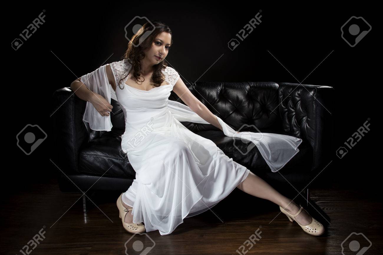 ブライダル レトロまたはアールデコのスタイルでシンプルだがエレガントな薄いウェディング ドレスの近代的なファッショントレンドをモデリングします 肩のスカーフとブライダル ガウンとソファの上の花嫁 の写真素材 画像素材 Image