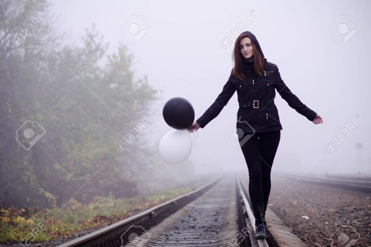 Lonely girl in black clothing walking on the rails in a fog. She holds a black and white balloon in her hand and it fly away. Concept of choice, target, good and evil, way in life - 138897308 Lonely girl in black clothing walking on the rails in a fog. She holds a black and white balloon in her hand and it fly away. Concept of choice, target, good and evil, way in life - 138897308