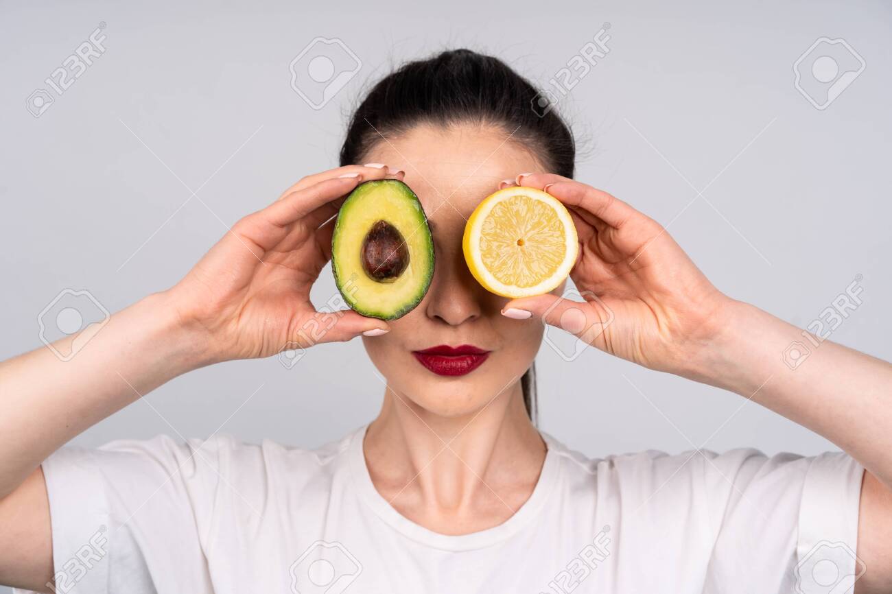 Girl in white shirt holds the halves of the avocado and orange right next to the eye, the concept of beauty and health - 149473378 Girl in white shirt holds the halves of the avocado and orange right next to the eye, the concept of beauty and health - 149473378
