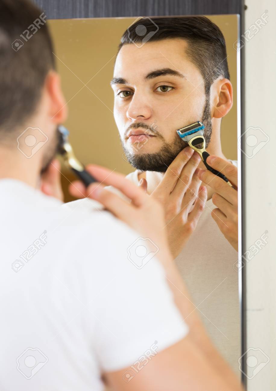 european man looking at mirror and shaving face with razor stock