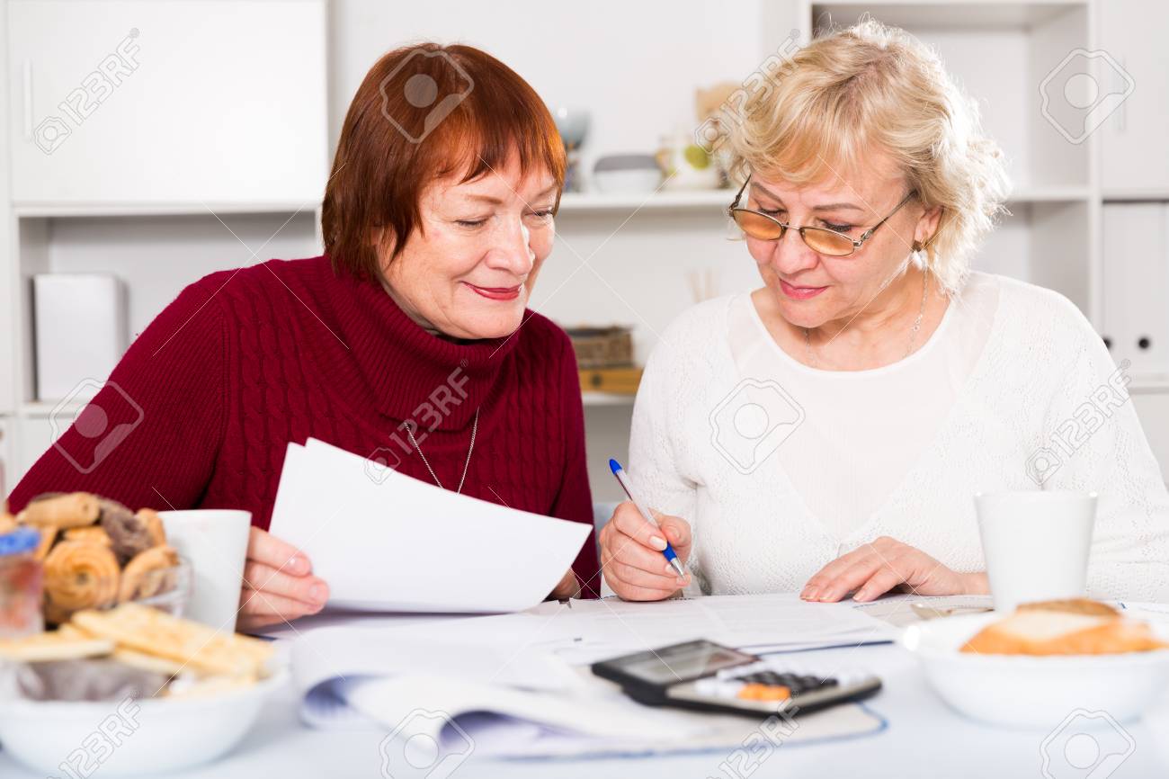 Stock Photo Two Positive Mature Women Considering Papers While Sitting On Kitchen Stock Photo Two Positive Mature Women Considering Papers While Sitting On Kitchen