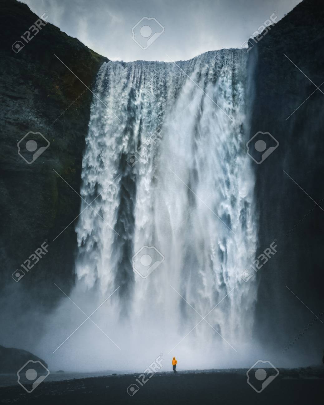 Panorama view of hiker in yellow rain jacket standing in front of gigantic Skogafoss waterfall on a moody day with dark clouds in summer, Skogar, Iceland - 113997483 Panorama view of hiker in yellow rain jacket standing in front of gigantic Skogafoss waterfall on a moody day with dark clouds in summer, Skogar, Iceland - 113997483