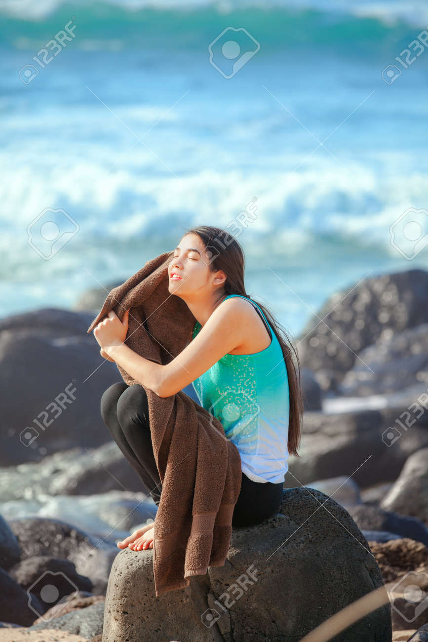 Belle Biracial Jeune Adolescent Fille Assise Sur La Plage Rocheuse à Hawaii Sécher Avec Une Grande Serviette