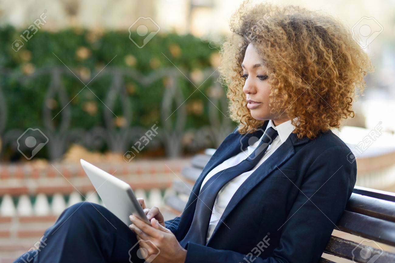 Beautiful black curly hair african woman using tablet computer on an urban bench. Businesswoman wearing suit with trousers and tie, afro hairstyle. - 55150666 Beautiful black curly hair african woman using tablet computer on an urban bench. Businesswoman wearing suit with trousers and tie, afro hairstyle. - 55150666