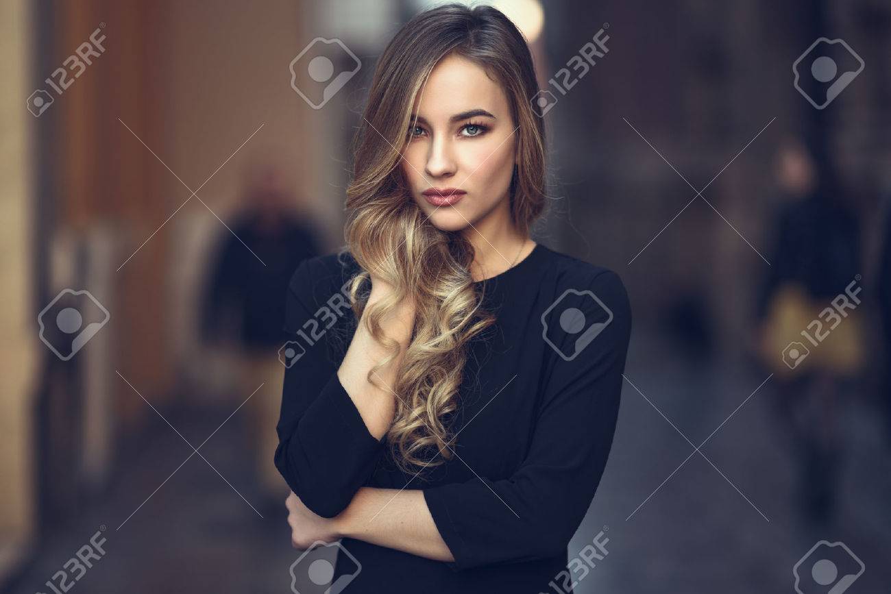 Blonde woman in urban background. Beautiful young girl wearing black elegant dress standing in the street. Pretty russian female with long wavy hair hairstyle and blue eyes. - 72165089 Blonde woman in urban background. Beautiful young girl wearing black elegant dress standing in the street. Pretty russian female with long wavy hair hairstyle and blue eyes. - 72165089