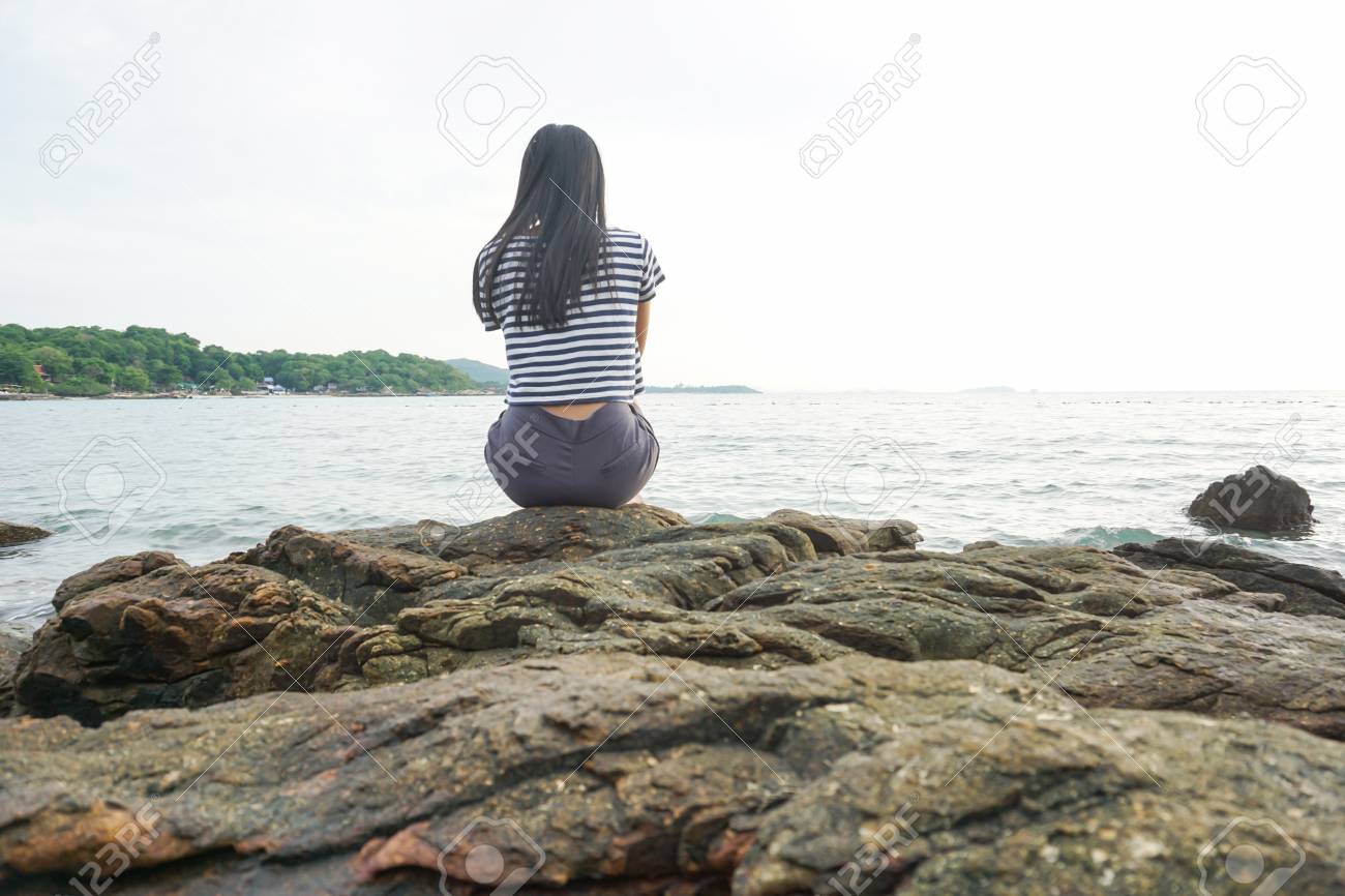 young woman sitting on rock facing the sea sad and depressed stock