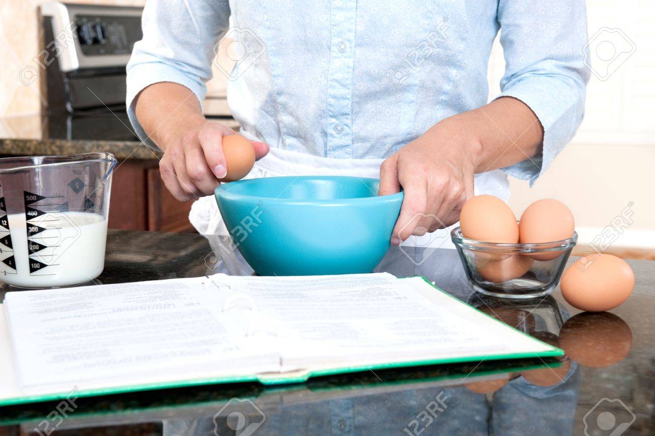 A homemaker cracks an egg into a mixing bowl while following the instructions of a cookbook. - 8430359 A homemaker cracks an egg into a mixing bowl while following the instructions of a cookbook. - 8430359
