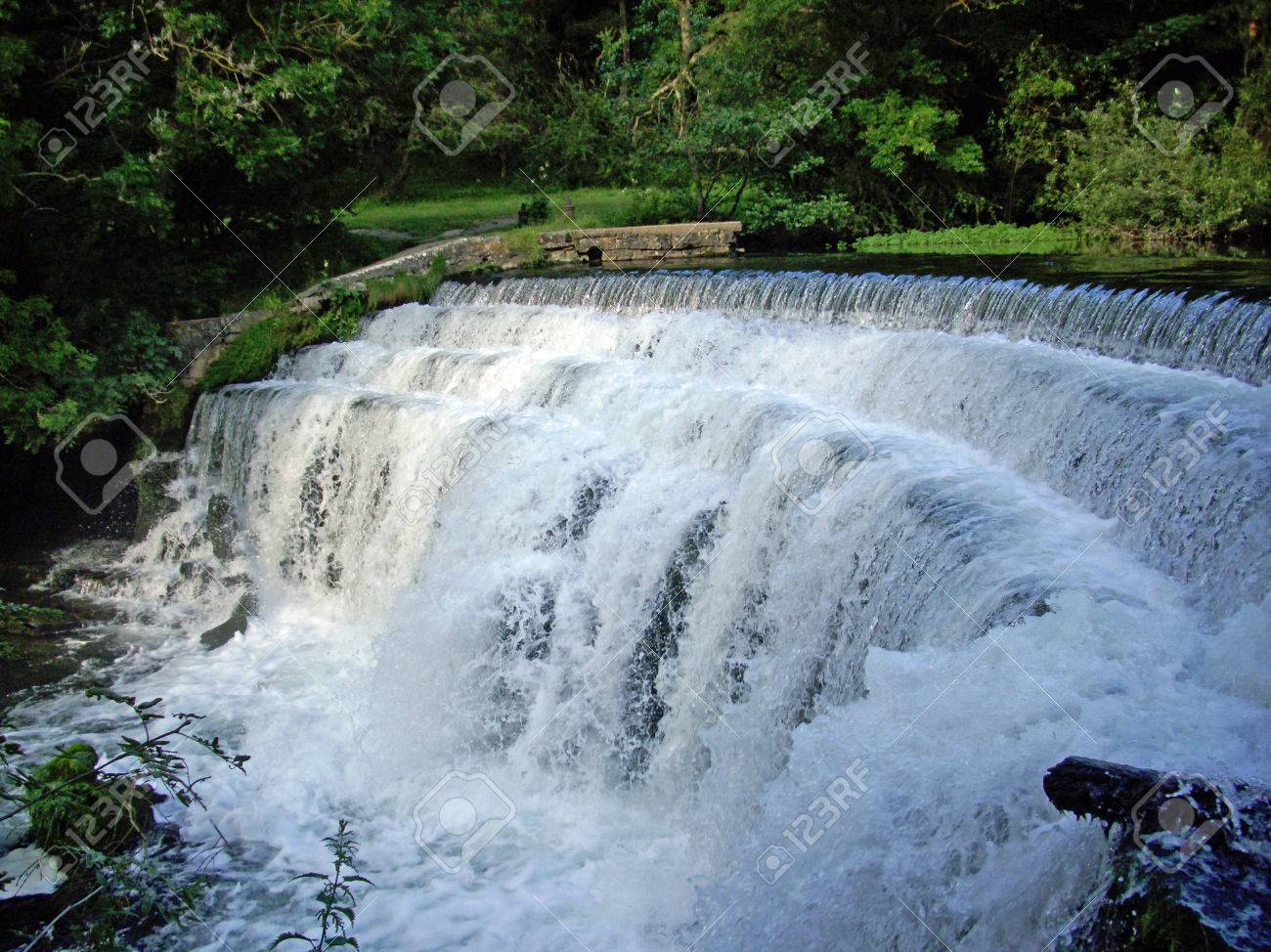 Gebogener Wasserfall Auf Einem Fluss In Der Flut Mit Weissem Schaum Und Spray Hintergrund Der Baume Und Einer Wiese Mit Einer Steinmauer Lizenzfreie Fotos Bilder Und Stock Fotografie Image 81265385
