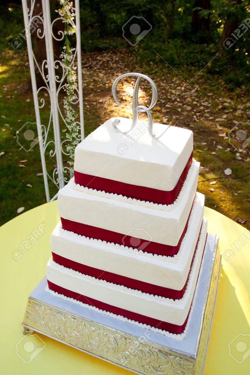 Un Gateau De Mariage Blanc Et Rouge Carre Avec Plusieurs Niveaux Et La Lettre R Sur Le Dessus Se Trouve A L Exterieur Sur Une Table Jaune Pour Une Reception De Mariage Banque