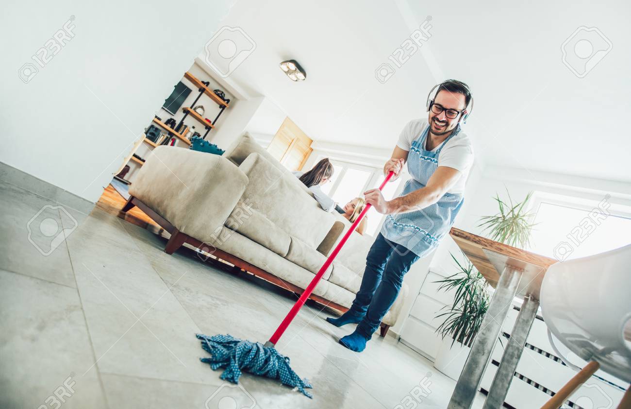 Husband housekeeping and cleaning concept. A man cleans the house, while women gossiping on the sofa - 121110942 Husband housekeeping and cleaning concept. A man cleans the house, while women gossiping on the sofa - 121110942