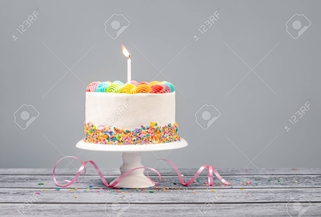 White Birthday cake with rainbow icing, colorful Sprinkles and one candle over a gray - 121745943 White Birthday cake with rainbow icing, colorful Sprinkles and one candle over a gray - 121745943