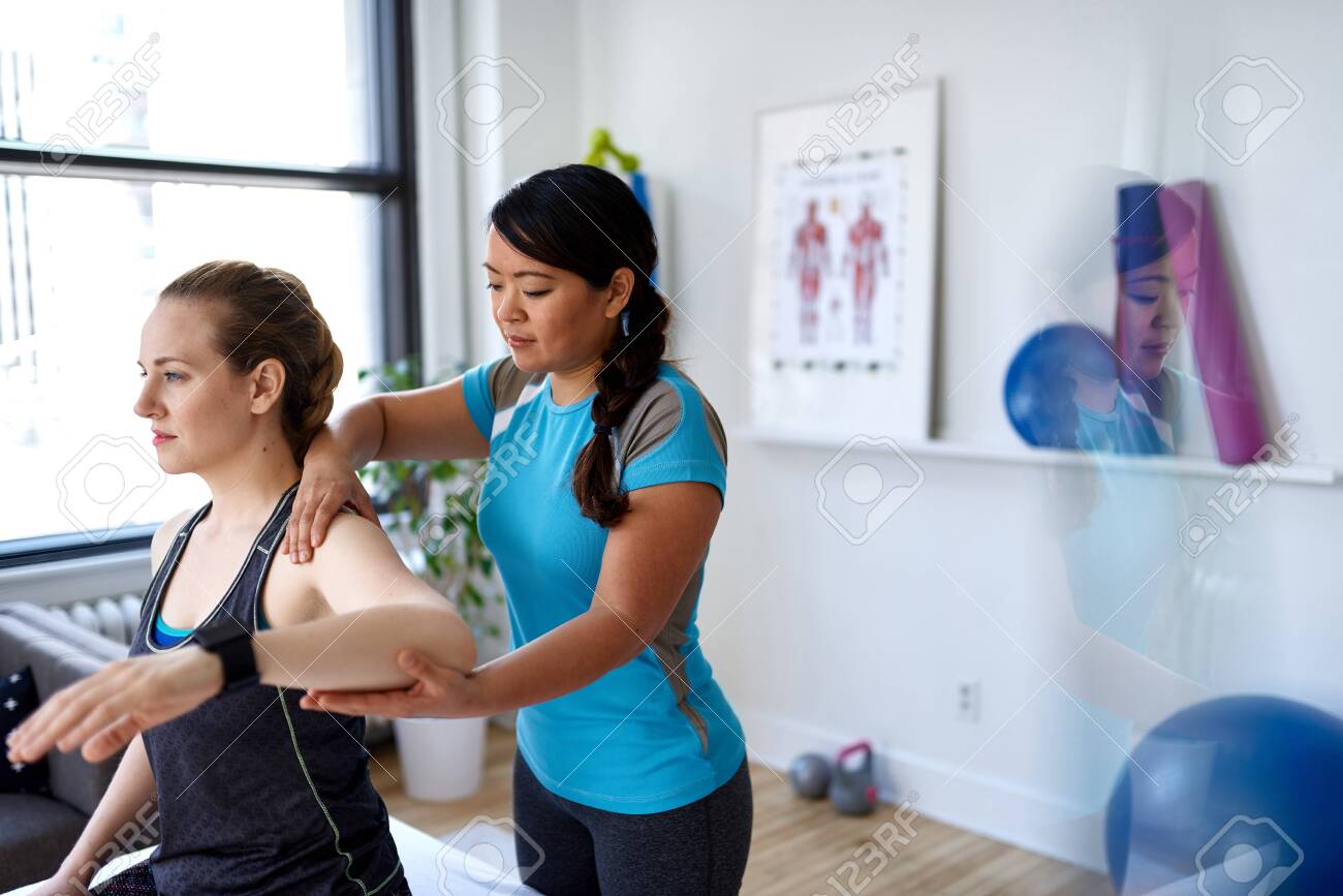 Chinese woman physiotherapy professional giving a treatment to an attractive blond client in a bright medical office - 131463653 Chinese woman physiotherapy professional giving a treatment to an attractive blond client in a bright medical office - 131463653