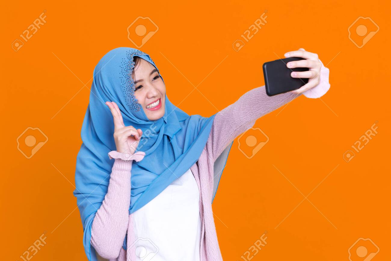 Happy Asian muslim woman taking selfie with smartphone isolated on color background - 149474077 Happy Asian muslim woman taking selfie with smartphone isolated on color background - 149474077