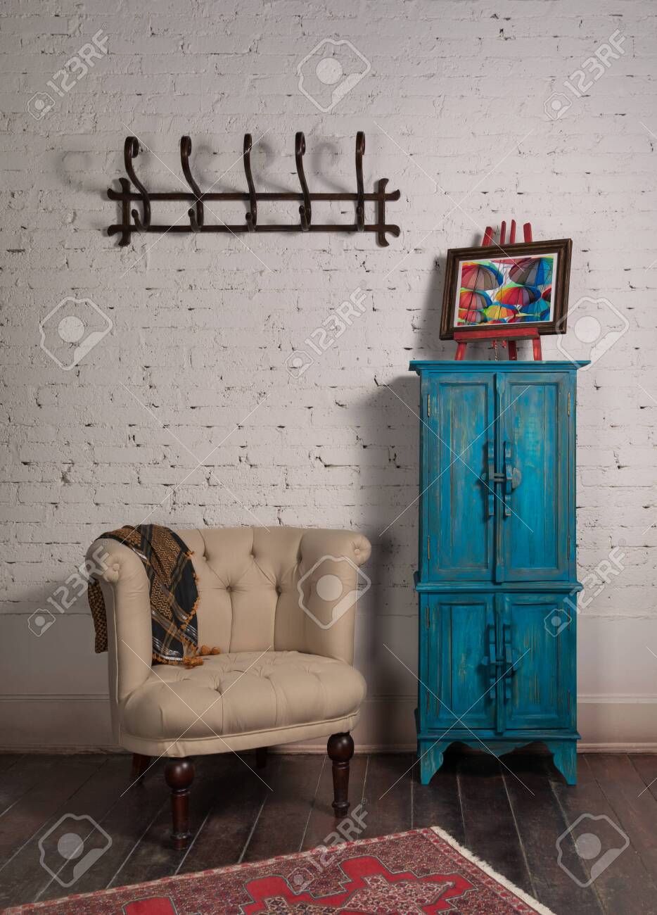 Classic beige armchair with wooden legs, vintage turquoise cupboard, wall hanger, and ornate scarf on white bricks wall and grunge wooden parquet with red decorated carpet - 151698897 Classic beige armchair with wooden legs, vintage turquoise cupboard, wall hanger, and ornate scarf on white bricks wall and grunge wooden parquet with red decorated carpet - 151698897