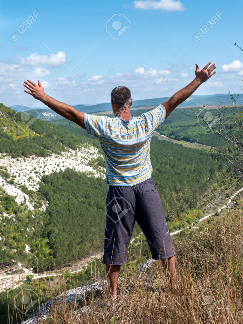 An adult man with his arms spread wide, seen from the back, stands on the edge of a precipice. - 155752173 An adult man with his arms spread wide, seen from the back, stands on the edge of a precipice. - 155752173