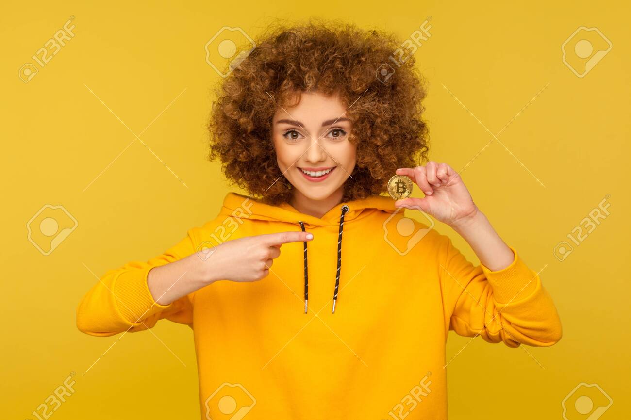 암호화폐, 디지털 화폐. 도시 스타일 후드티를 입은 행복한 곱슬머리 여성의 초상화는 황금색 비트코인을 보여주고 웃고, Btc 동전을  가리키고 있습니다. 노란색 배경에 고립 된 실내 스튜디오 촬영 로열티 무료 사진, 그림, 이미지 그리고