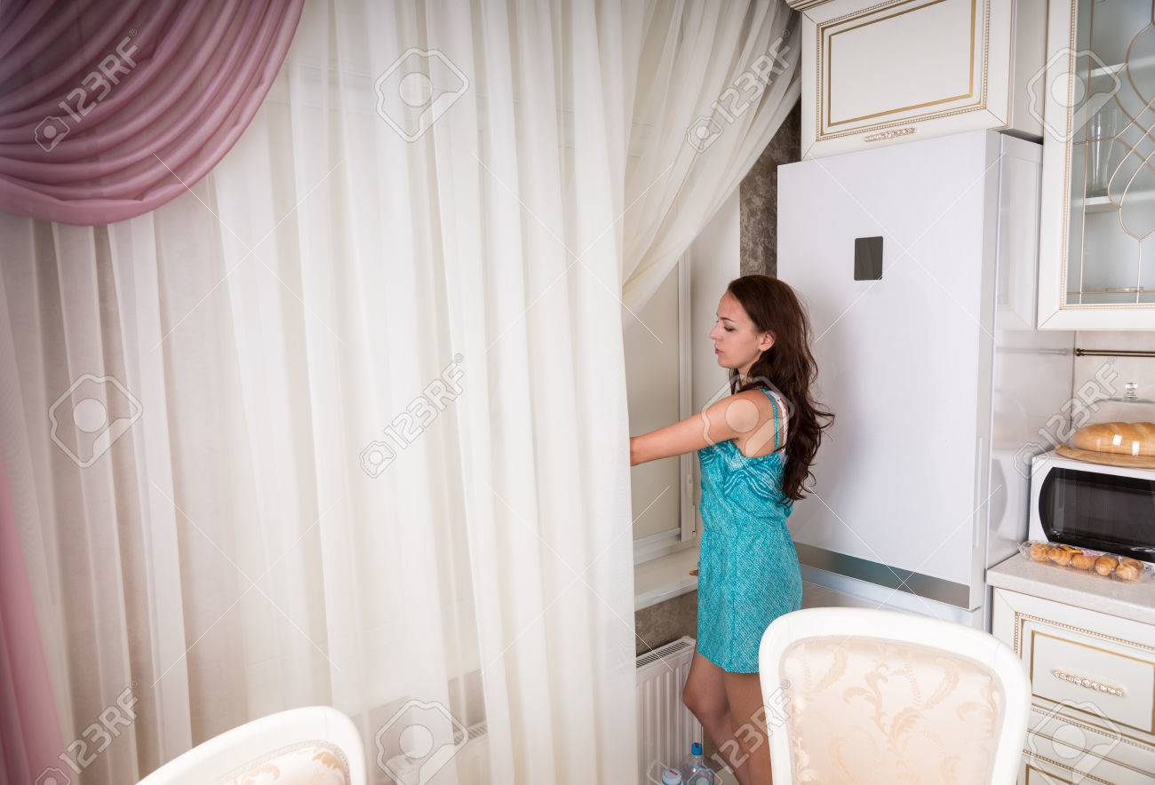 台所の窓の外を見て若いブルネットの女性期待し待っている 女性を脇に押しカーテンから外を参照してください の写真素材 画像素材 Image