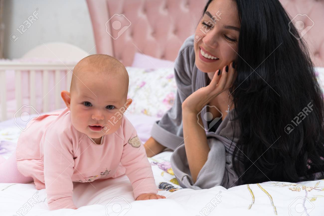 Young mother with long black hair with her young baby girl in bed looking at her daughter and smiling the child is dressed in pink dress standing on all